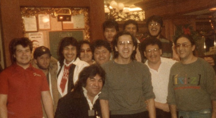 Image of a group in Gio's Bar, Winnipeg, Manitoba. Pictured: Perry Lajoie, Rocky Laroque, Clare Burns, Leroy Jebb, Roger Armitte, Peetanacoot Nenawekapo, Winston Thompson, Albert McLeod, Micky Morin, Leonard Carriere, John Burrell, Victor Apetagon (back, at the far right with glasses and a beard) : [between 1986 and 1989] : photograph : col.